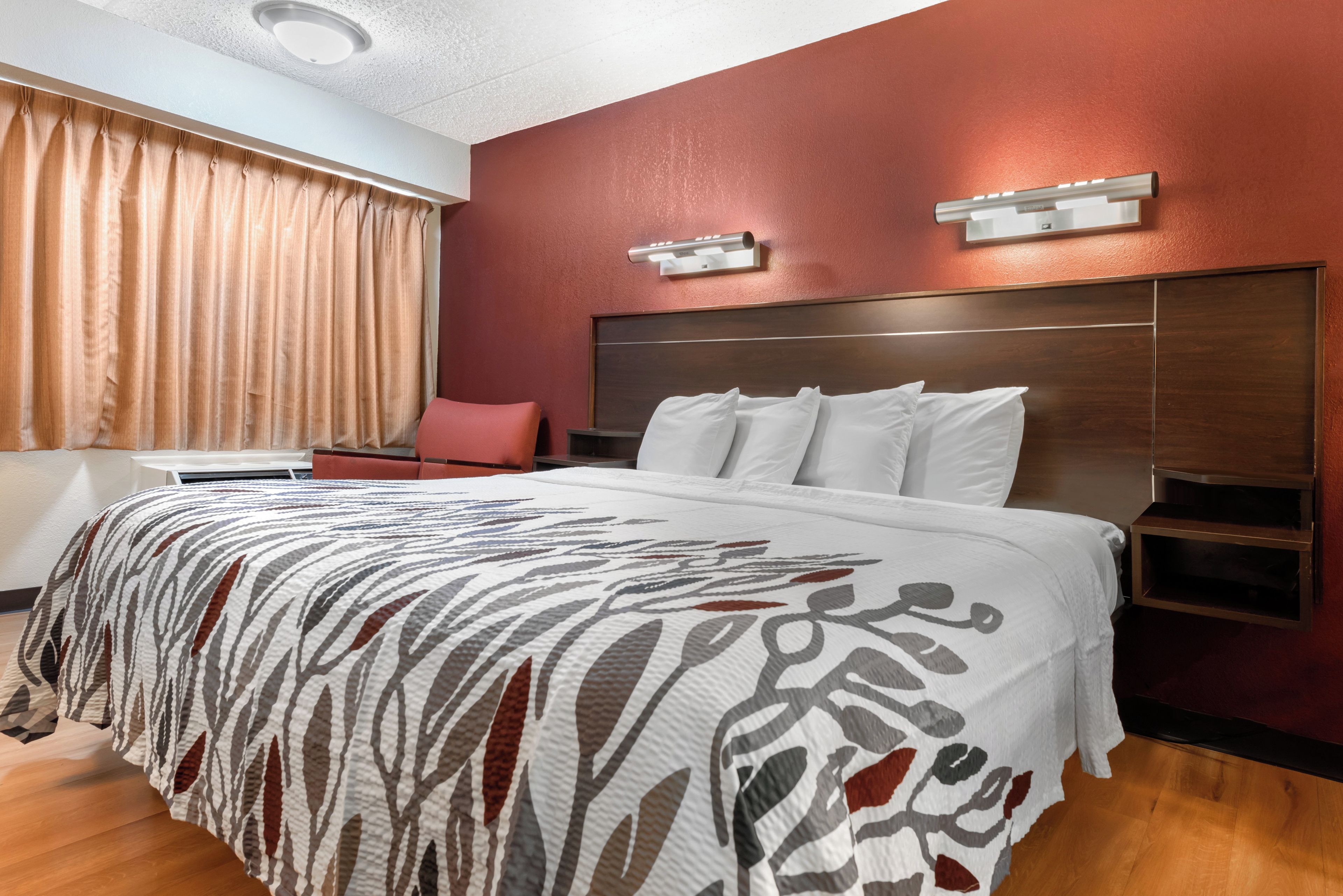 A hotel room with a large bed, a wooden headboard, a nightstand, a red chair, and a window with curtains.