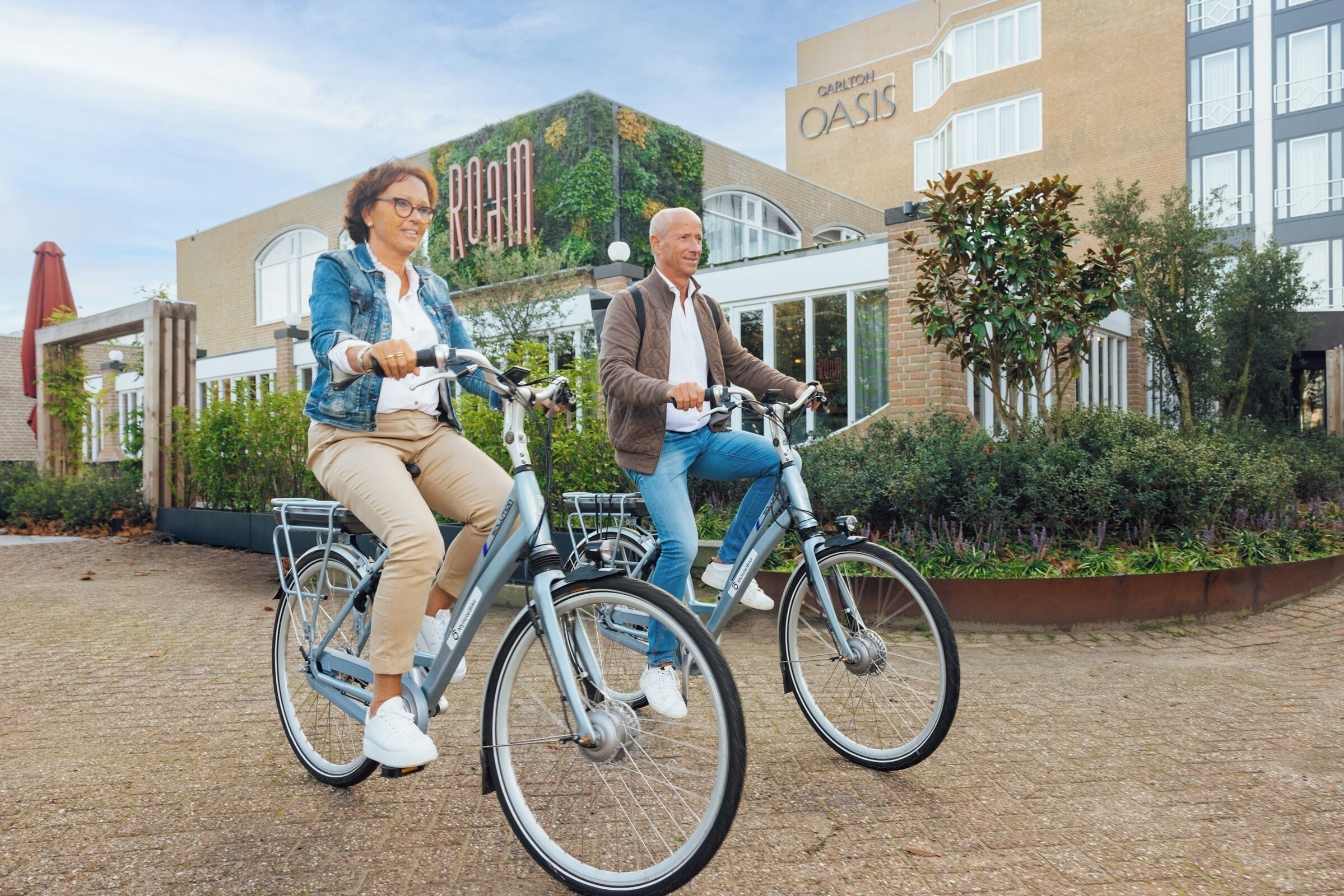 Dos personas montando en bicicleta frente a un edificio con un letrero que dice "Carlton Oasis".