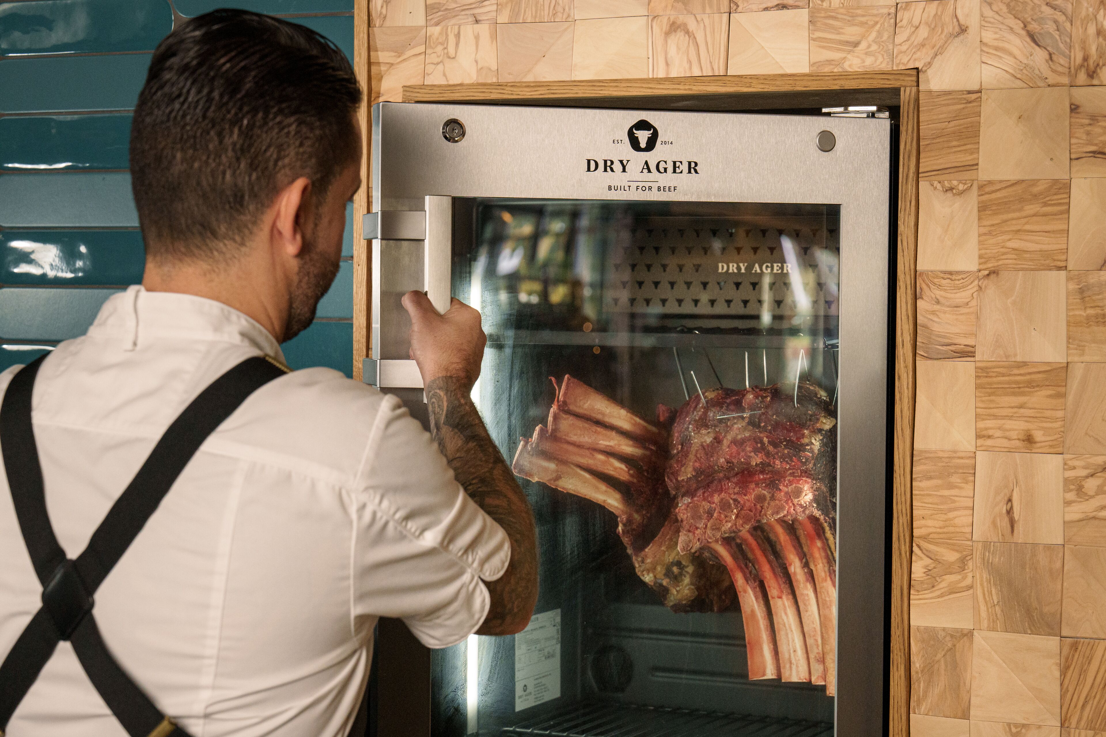 Eine Person in weißem Hemd und schwarzer Schürze öffnet ein Dry Ager Gerät zum Braten von Fleisch.