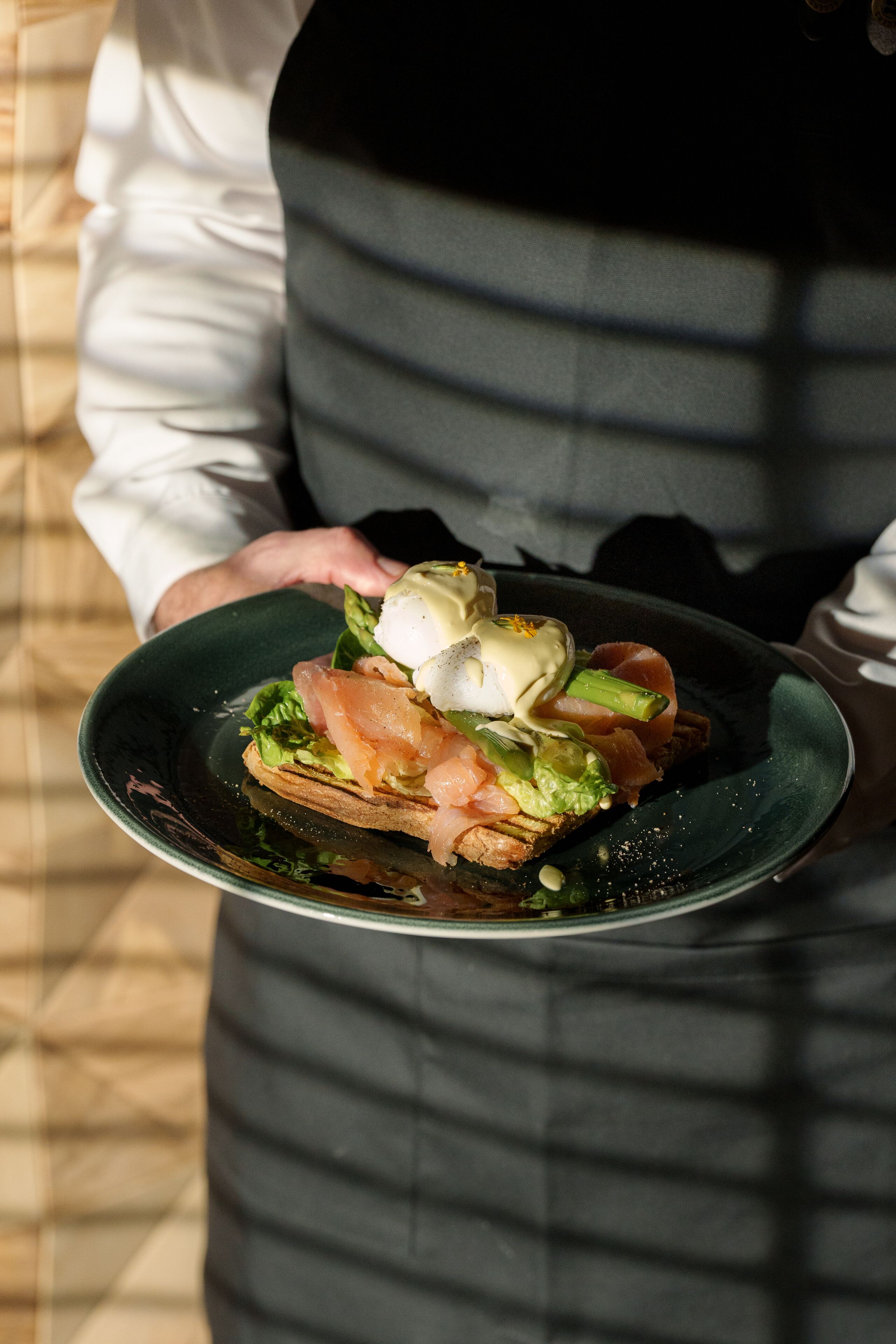 A person holding a plate with a sandwich, greens, and a poached egg.