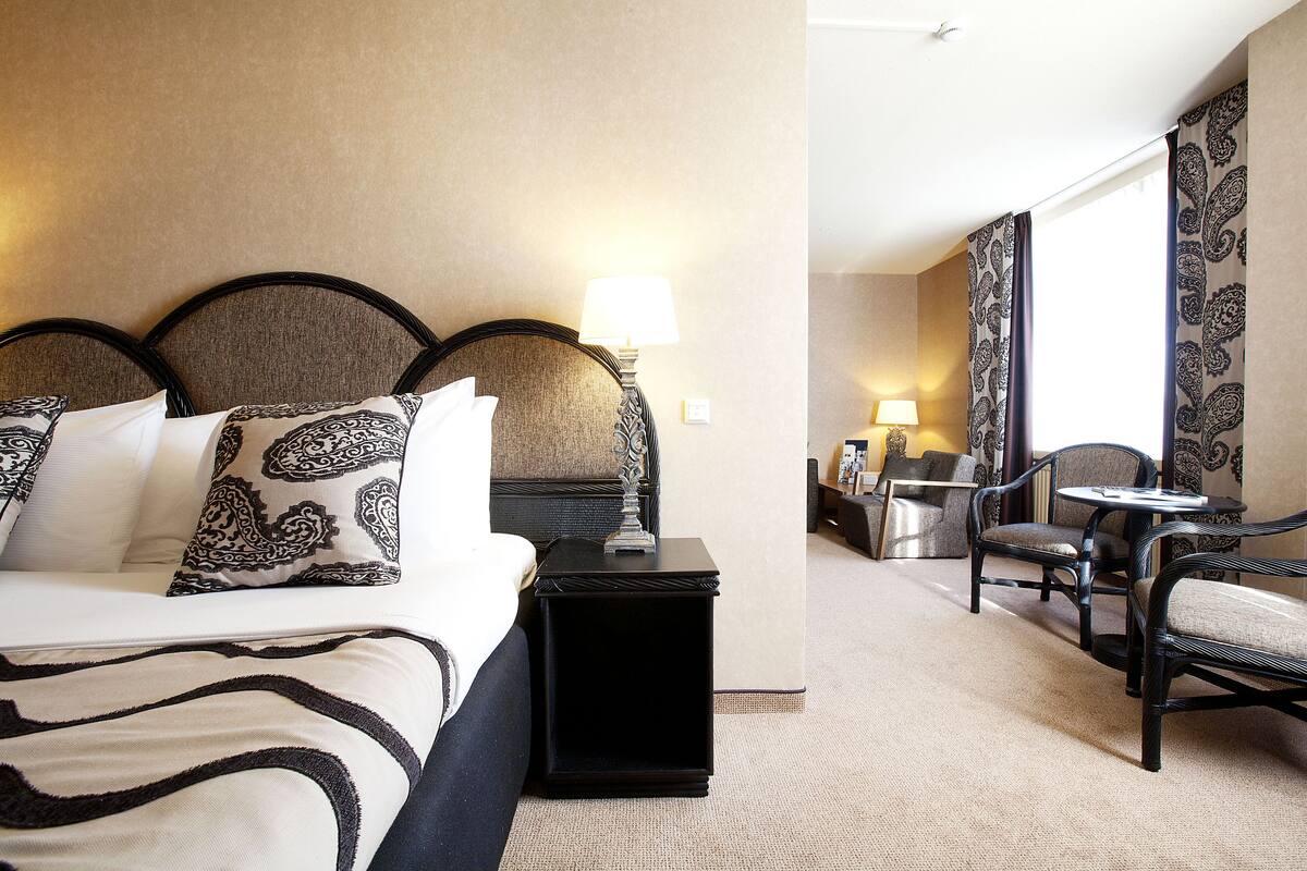 A hotel room with a large bed, a bedside table with a lamp, a small table with a chair, and a view of the hallway.