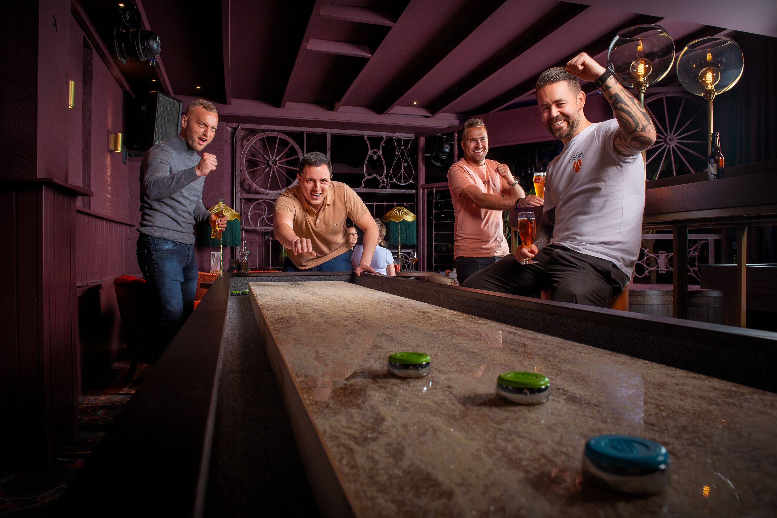 Ein Billardtisch mit vier Spielern, eine Bar mit Hockern und ein dekoratives Rad im Hintergrund.