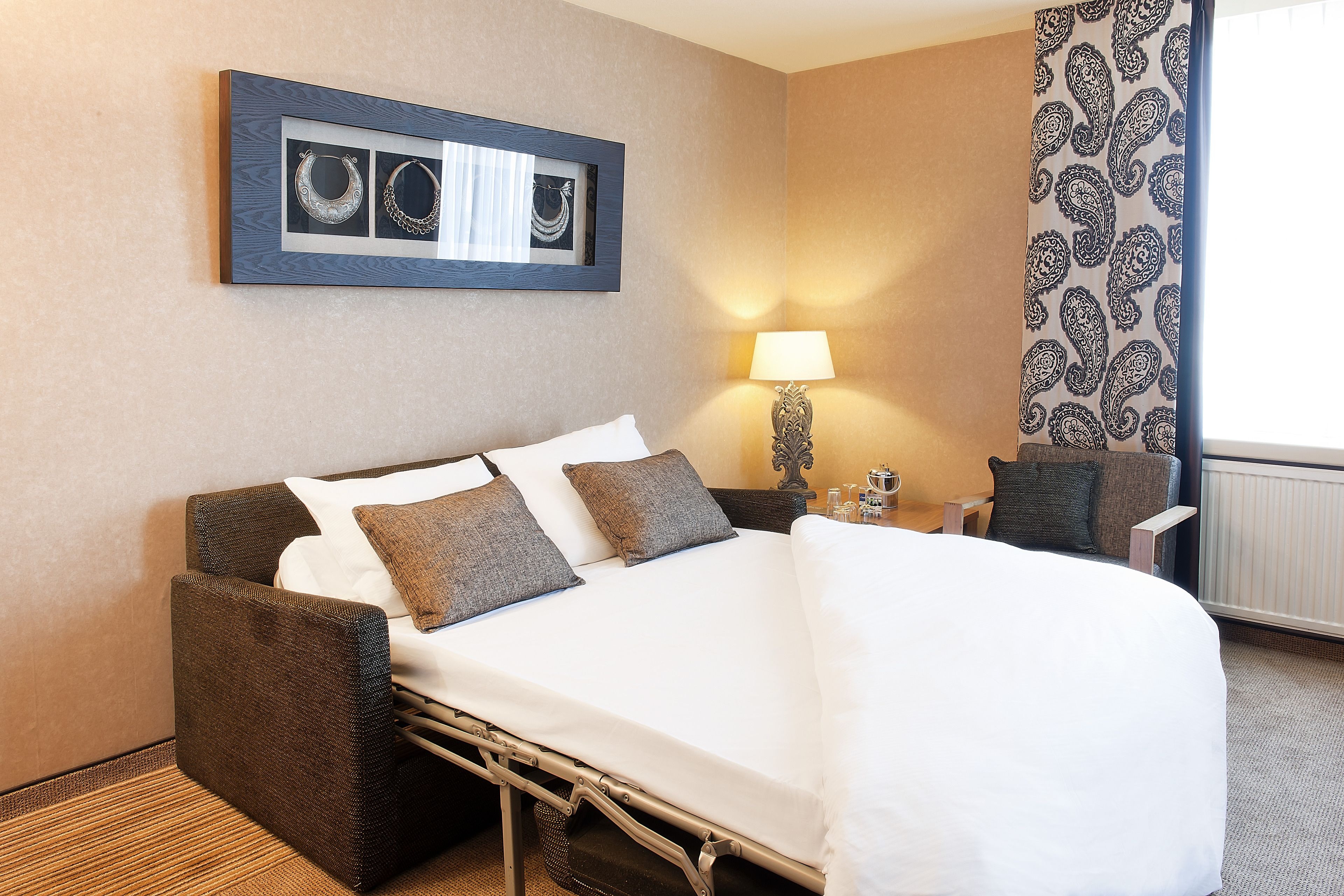 A hotel room with a bed, pillows, a nightstand, a lamp, and a framed picture on the wall.