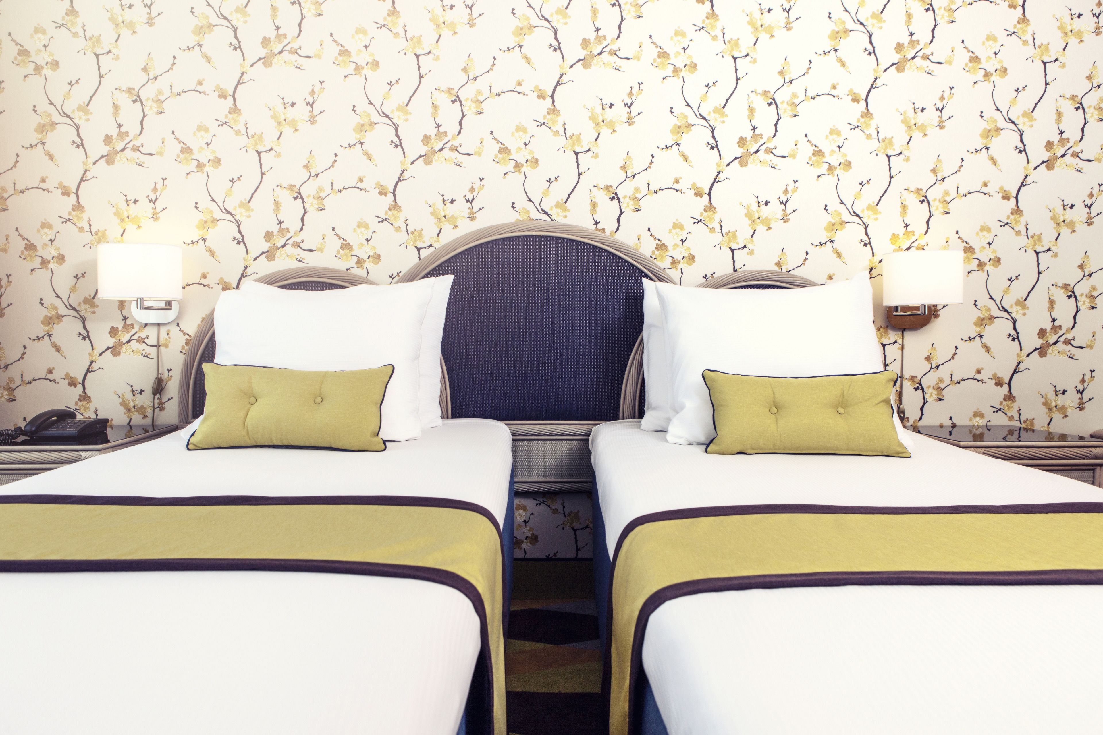 A hotel room with two beds, a headboard, bedside lamps, and a floral wallpaper.