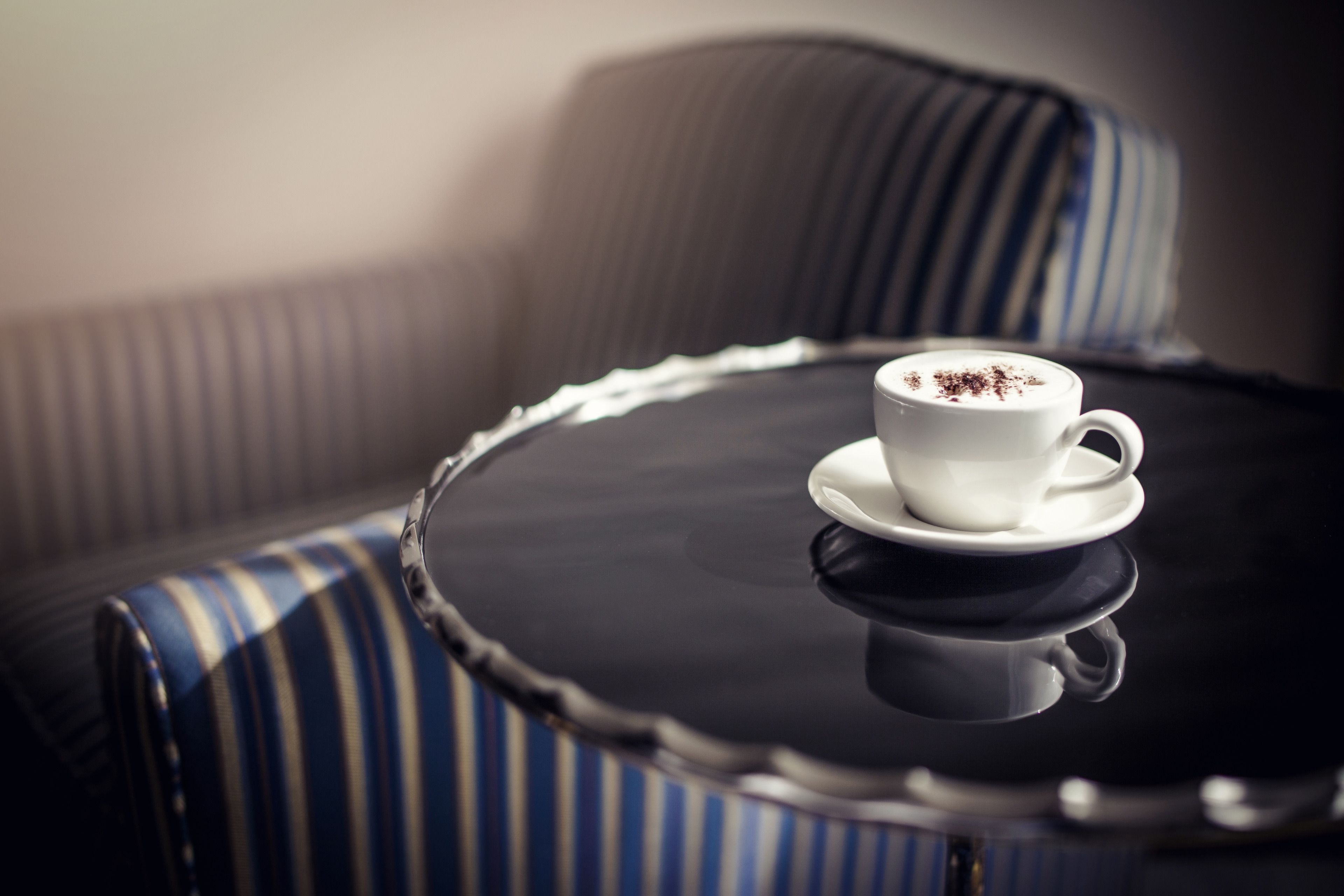 A white cup of coffee on a saucer on a round table with a black reflective surface, placed on a striped sofa.