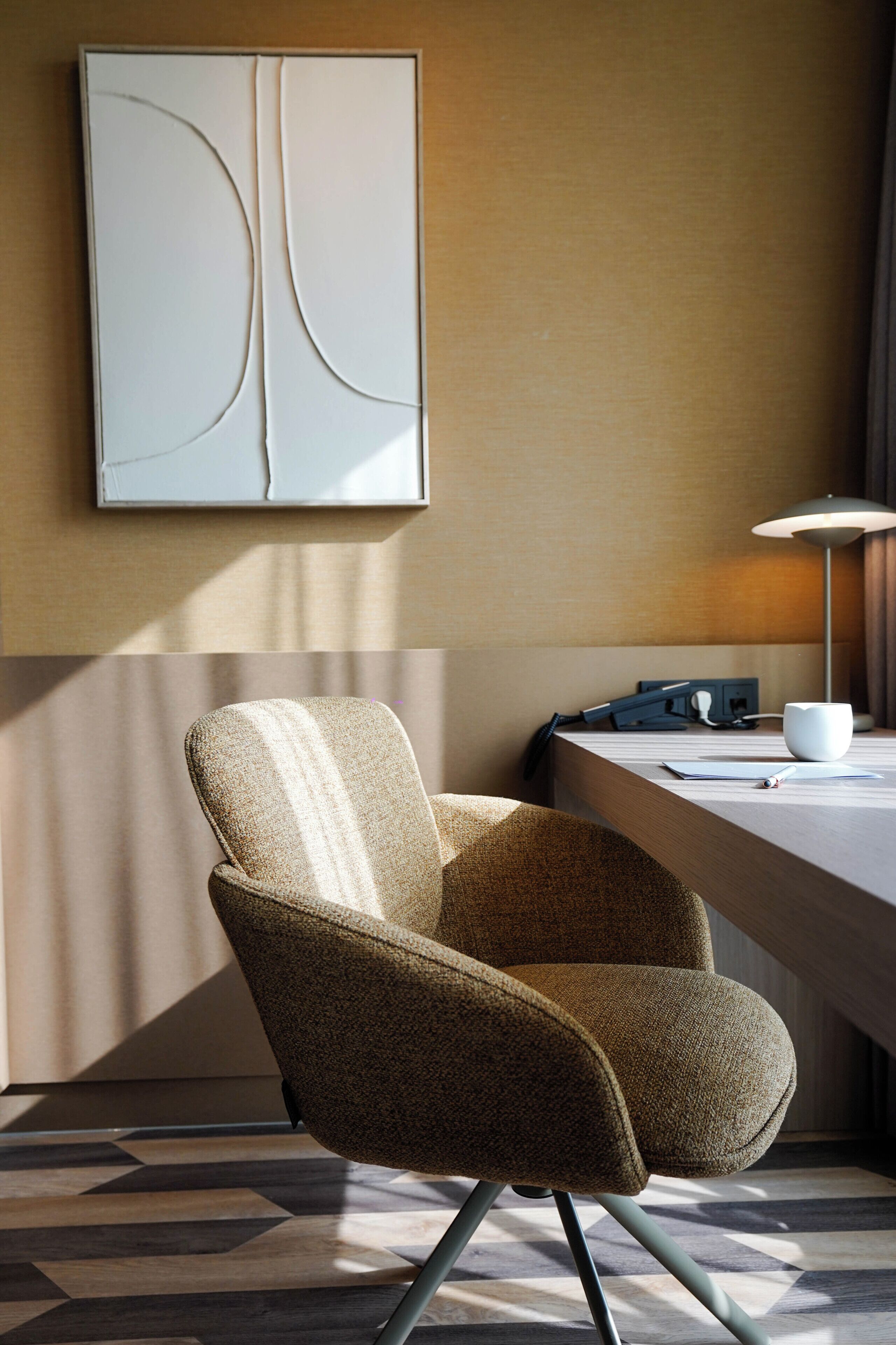 A modern hotel room with a wooden desk, a lamp, a chair, and a large abstract painting on the wall.