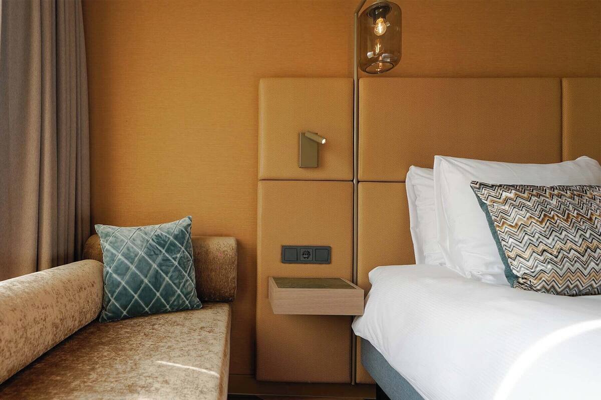 A hotel room with a bed, a bedside table, a lamp, and a sofa.