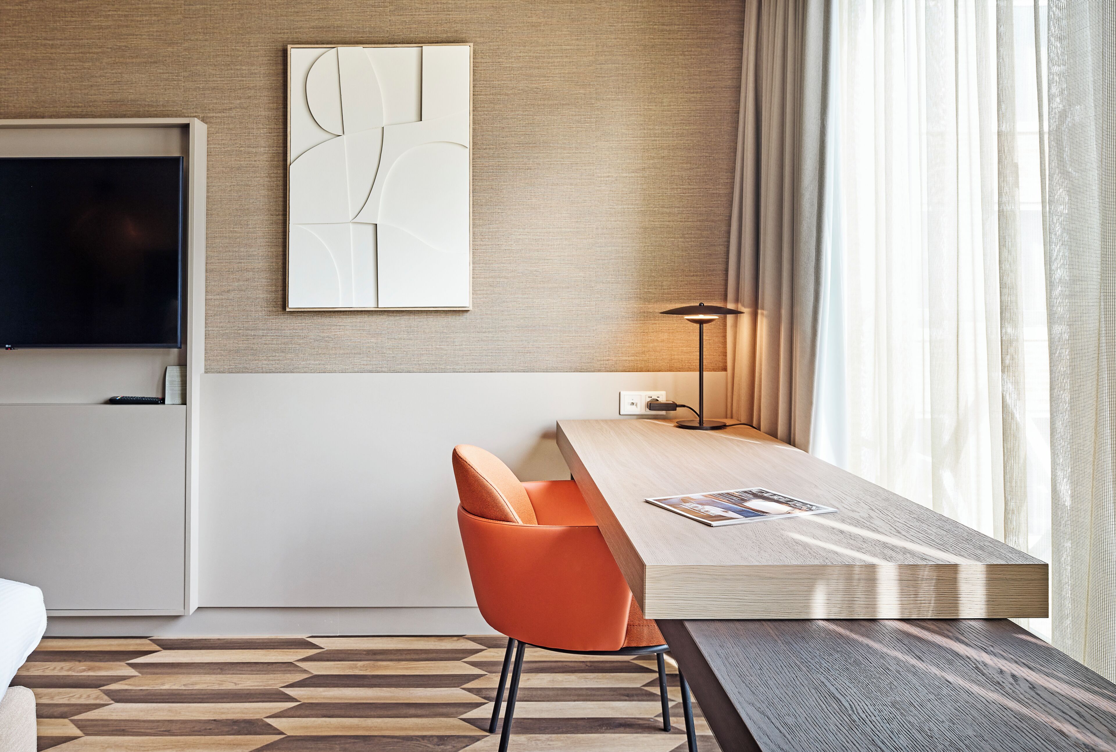 Une chambre d’hôtel moderne dotée d’un téléviseur à écran plat, d’une table à manger en bois, d’une chaise orange et d’une grande fenêtre ornée de rideaux légers.