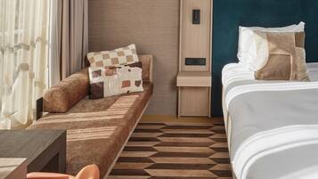 A hotel room with a sofa bed, a bedside table, a small desk, and a bed with pillows.