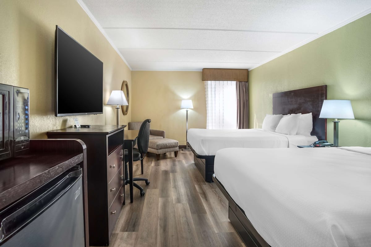 A hotel room with a flat-screen TV, a desk with a chair, two beds, a small table, and a lamp.