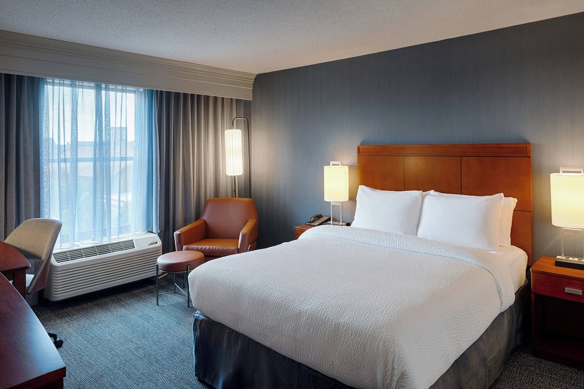 A hotel room with a large bed, two bedside lamps, a desk with a chair, a desk lamp, a window with curtains, and a wall-mounted air conditioner.