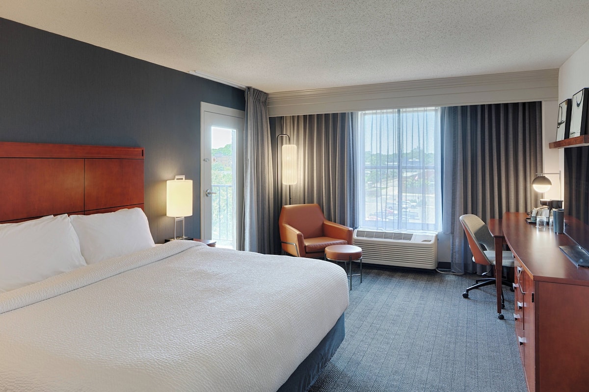 A hotel room with a large bed, a desk, a chair, and a view of the outdoors.
