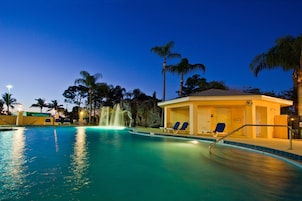 A swimming pool with a waterfall feature, lounge chairs, and palm trees.