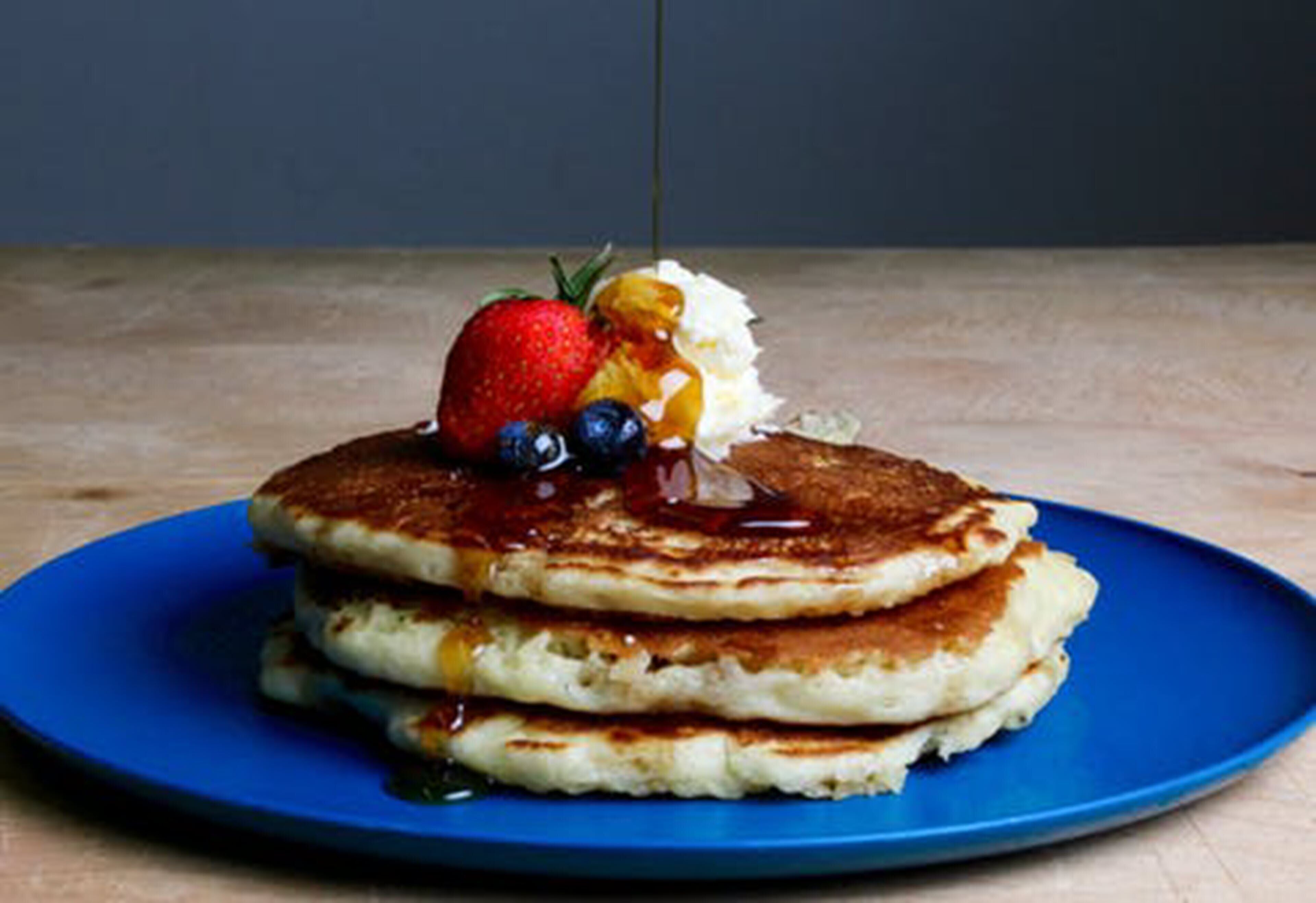 Une pile de crêpes sur une assiette bleue, nappées de sirop, garnies d’une fraise, de myrtilles et d’une quenelle de crème fouettée.