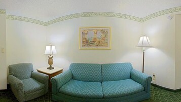 A room with a patterned sofa, a chair, a small table with a lamp, and a framed picture on the wall.