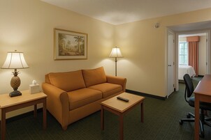 Une chambre d’hôtel avec un canapé, deux tables, une télécommande, une lampe, un tableau et une porte donnant sur une salle de bain.