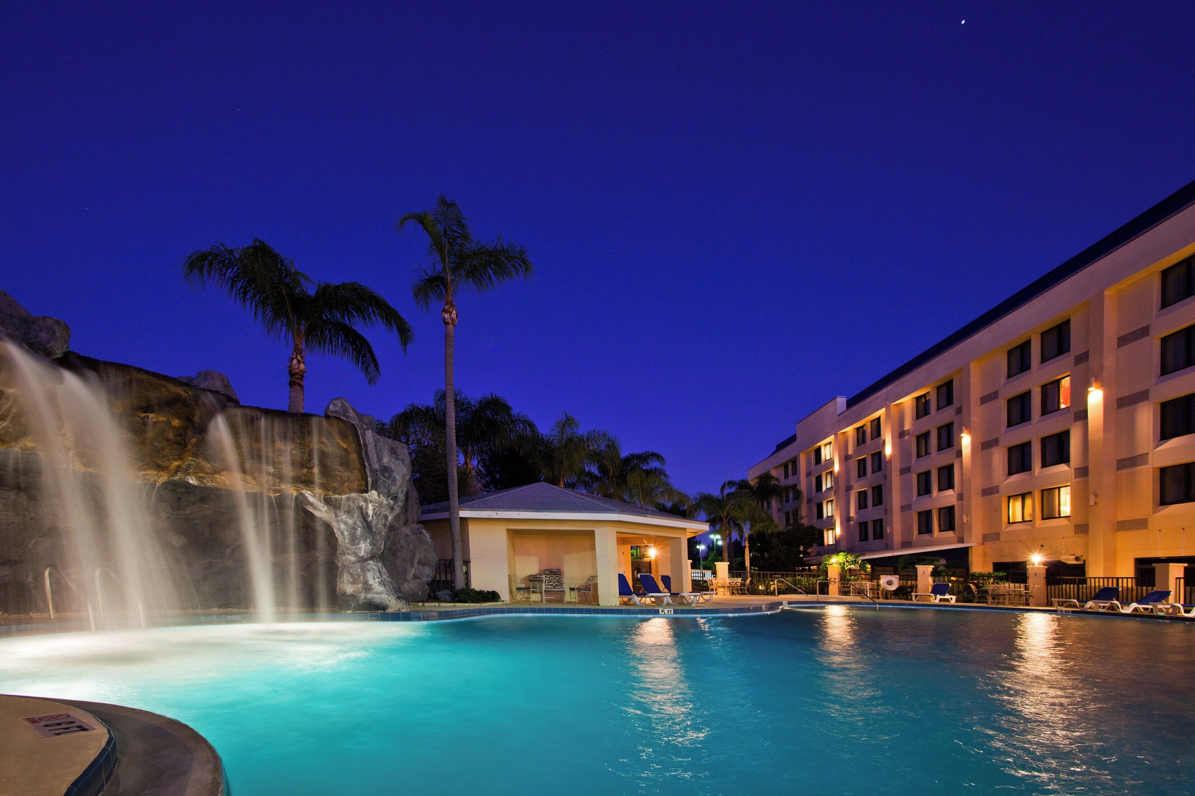 Piscina de hotel con cascada, palmeras y un edificio de varios pisos con balcones.