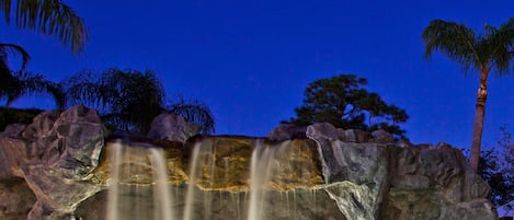 A nighttime view of a pool with a waterfall feature, surrounded by rocks and palm trees.