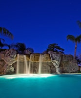 Vista nocturna de una piscina con cascada, rodeada de rocas y palmeras.