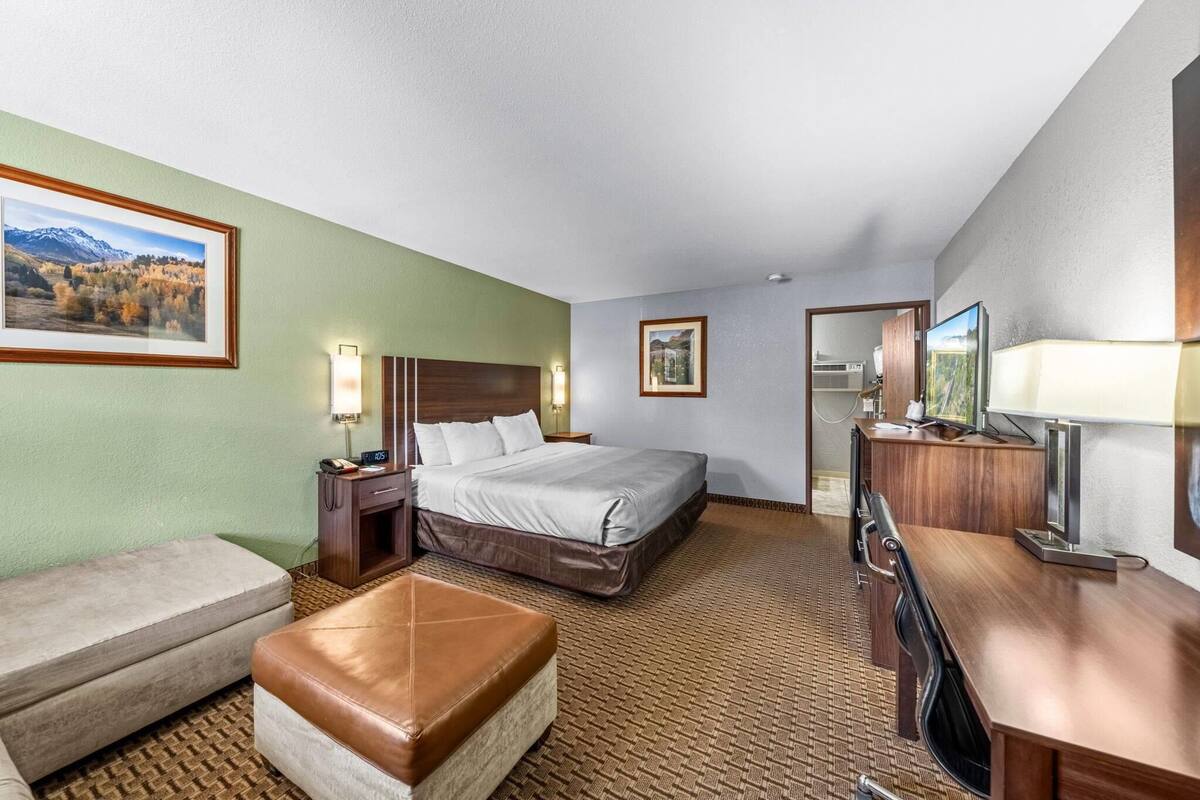 A hotel room with a bed, a desk, a chair, a bench, and a TV.