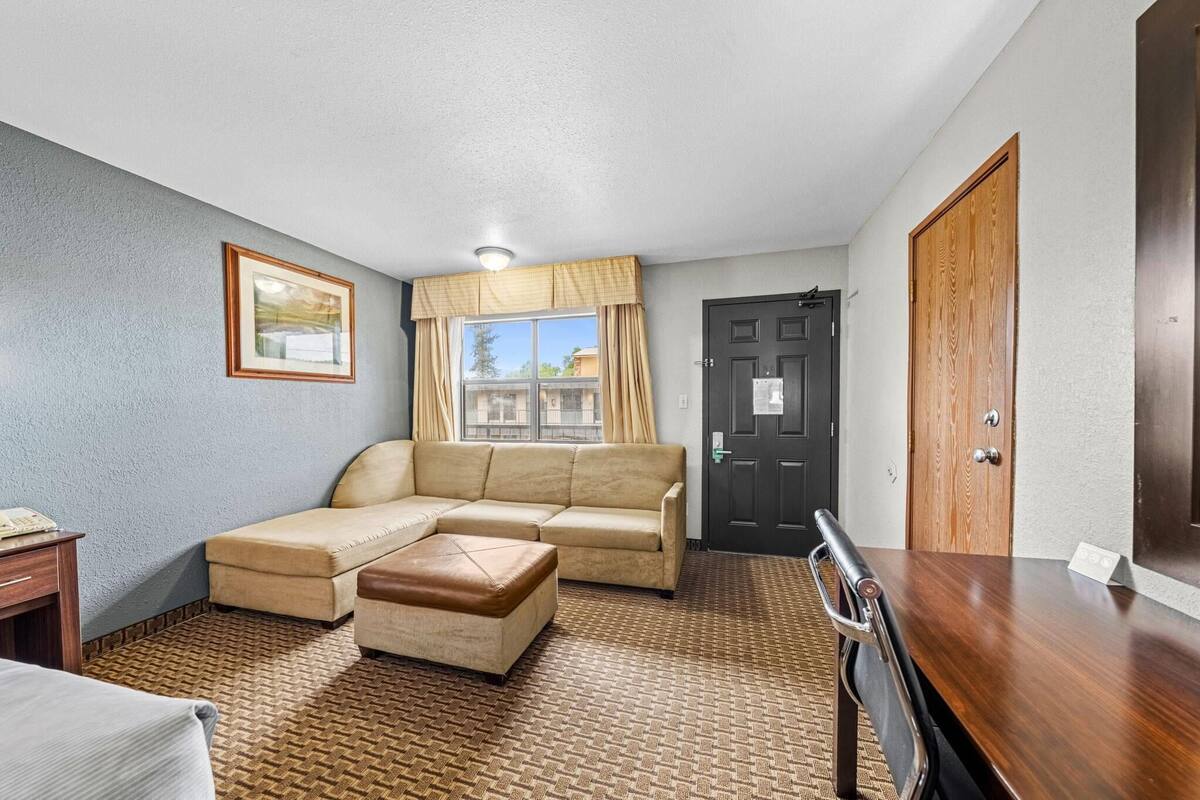 A hotel room with a beige sectional sofa, a wooden dining table, a small wooden desk, and a window with curtains.