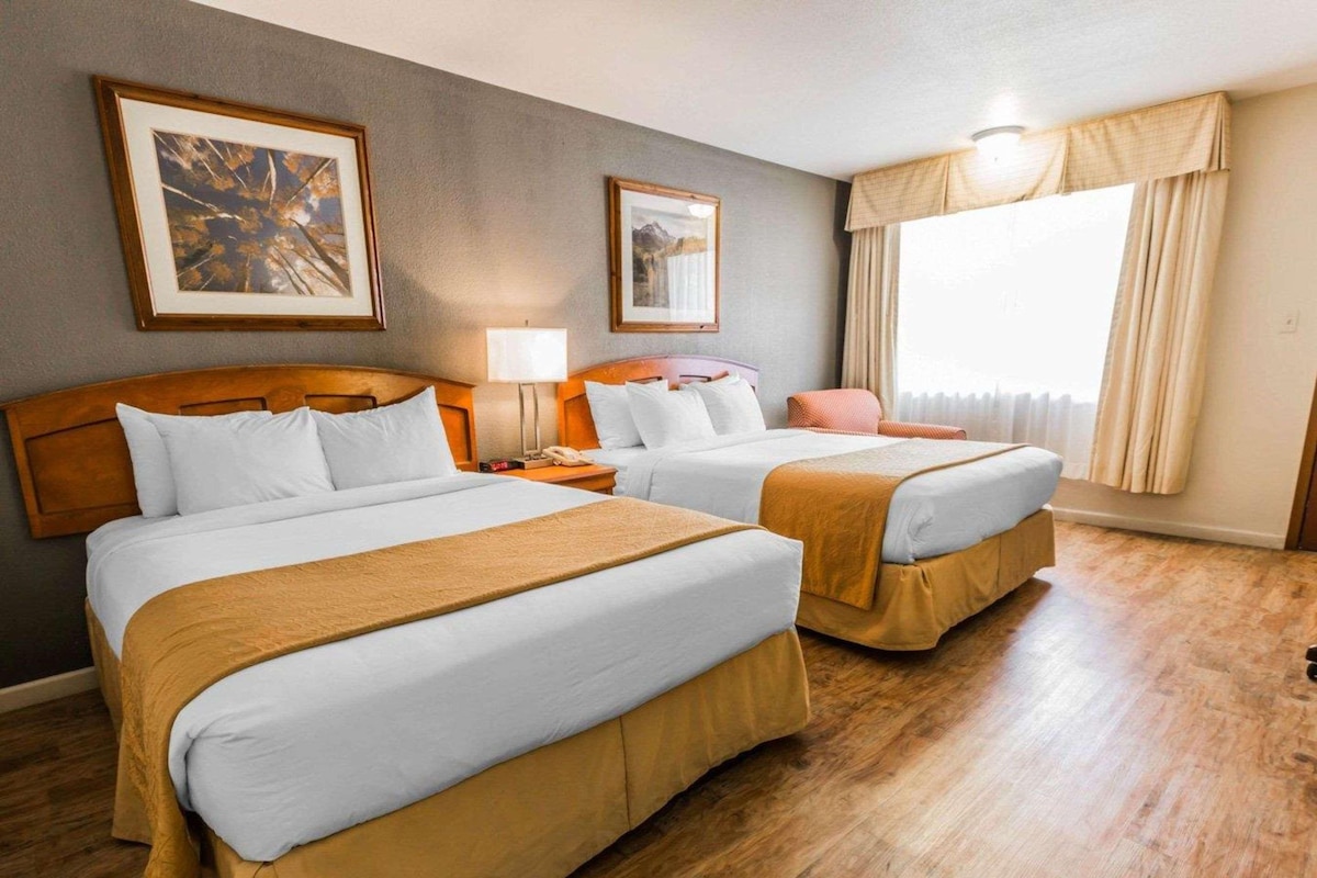 A hotel room with two beds, wooden headboards, a large window with curtains, and two framed pictures on the wall.