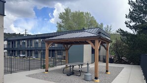 A covered outdoor seating area with a table and chairs, surrounded by a fence and a building in the background.