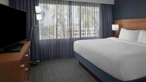 Una habitación de hotel con una cama grande, un televisor de pantalla plana, un escritorio con lámpara y vistas a palmeras desde la ventana.