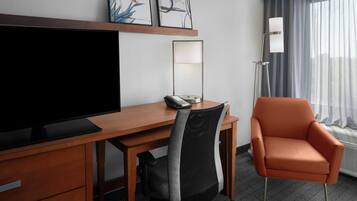 A hotel room with a wooden desk, a television, a chair, a desk lamp, and artwork on the wall.