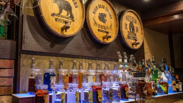 A bar area with wooden barrels labeled "Wild Bear Tavern Whiskey" and a variety of liquor bottles.