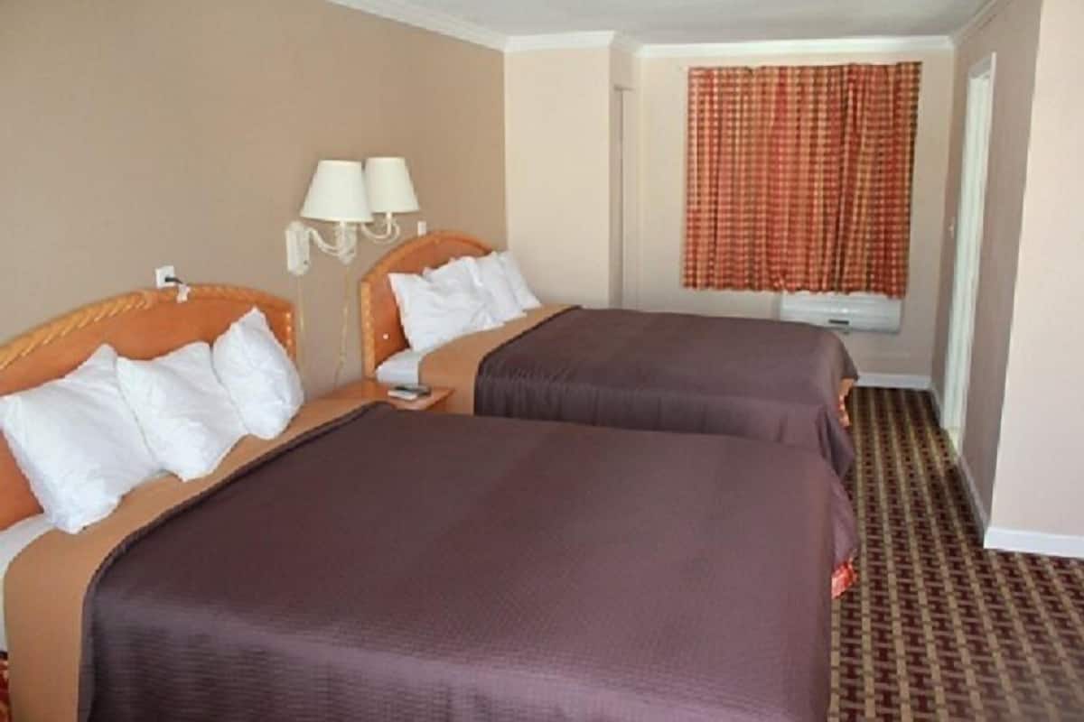 A hotel room with two beds, a window with curtains, a wall-mounted lamp, and a patterned carpet.