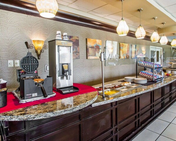 A breakfast bar with waffle maker, coffee machine, and a variety of food items.