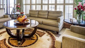 A hotel lobby with a sofa, a round table with a vase, and large windows with blinds.