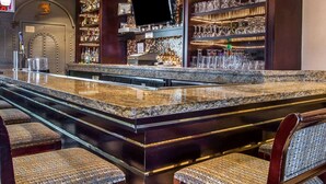 A bar area with a granite countertop, multiple bar stools, a television, and shelves stocked with bottles and glasses.