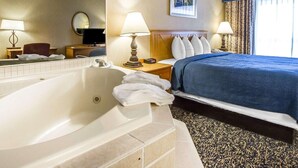 A hotel room with a large bathtub, a bed with a blue bedspread, a wooden headboard, a bedside table with a lamp, and a framed picture on the wall.