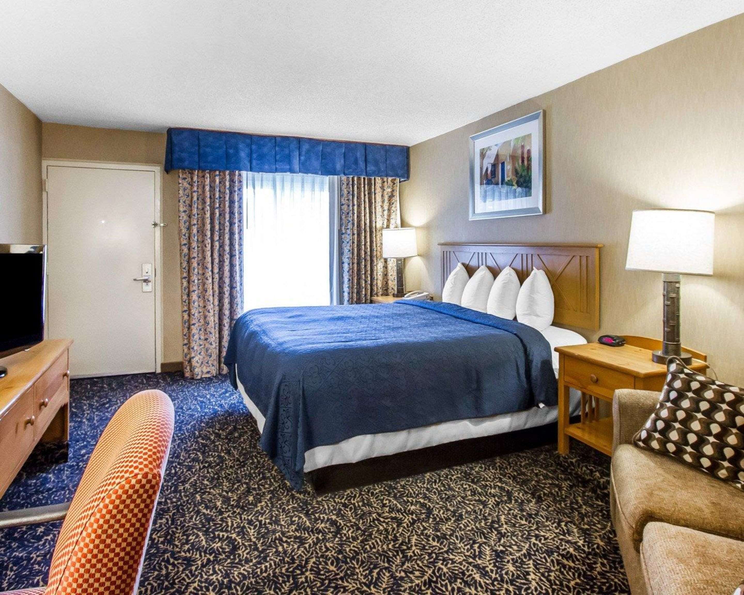 A hotel room with a large bed, a desk with a lamp, a chair, and a television.