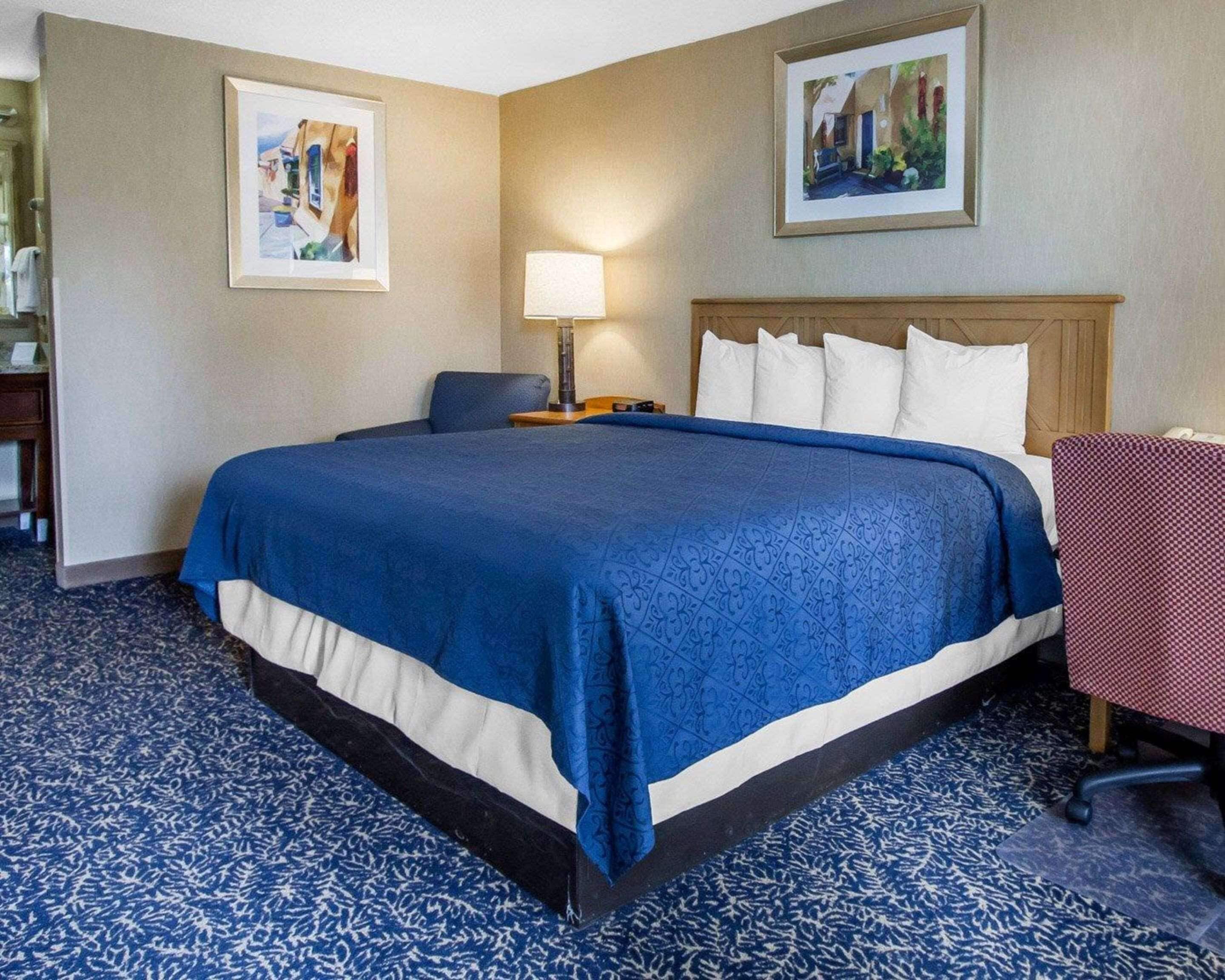 A hotel room with a large bed, a desk with a lamp, and two framed pictures on the walls.