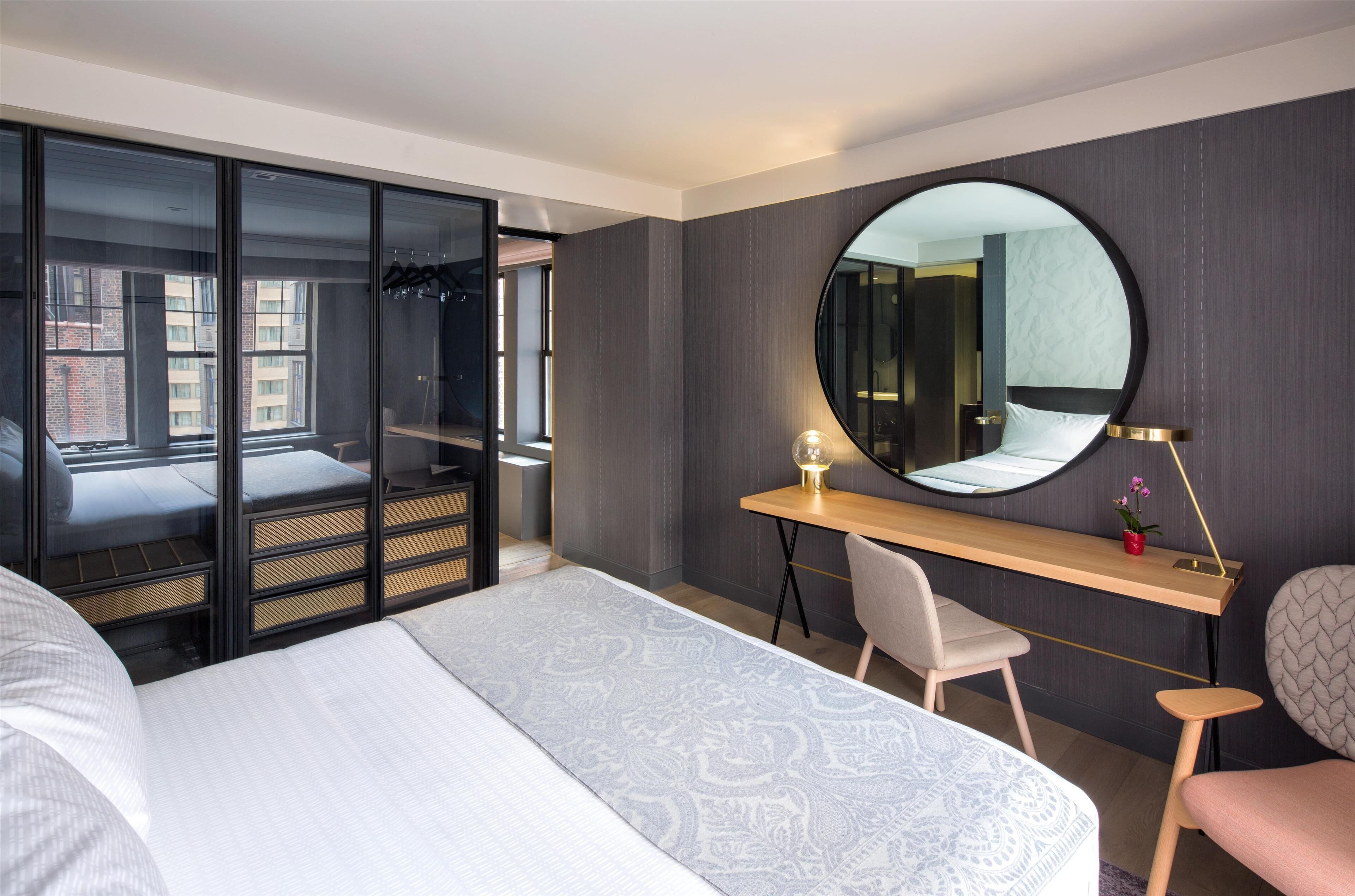 A modern hotel room with a large bed, a desk with a chair, a round mirror, and a wardrobe with sliding doors.