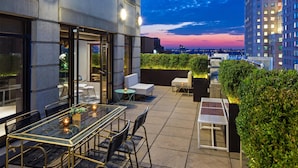 An outdoor terrace with a glass table, chairs, and a seating area with cushions. There are plants and a view of the city skyline at dusk.