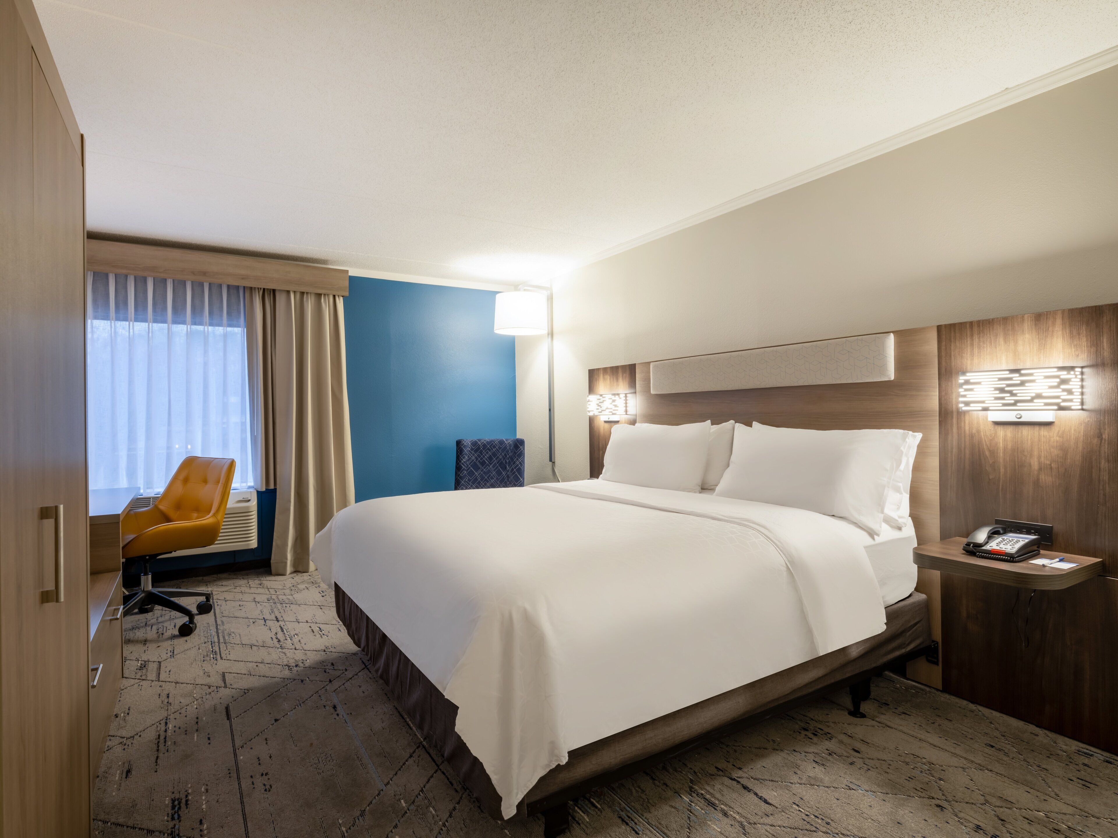 A hotel room with a large bed, a desk with a chair, a bedside table with a phone, and a window with curtains.