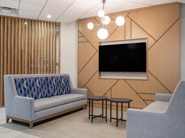Een moderne woonkamer met een grijze bank, een decoratief kussen, een flatscreen televisie bevestigd aan een houten wand en een kleine tafel.