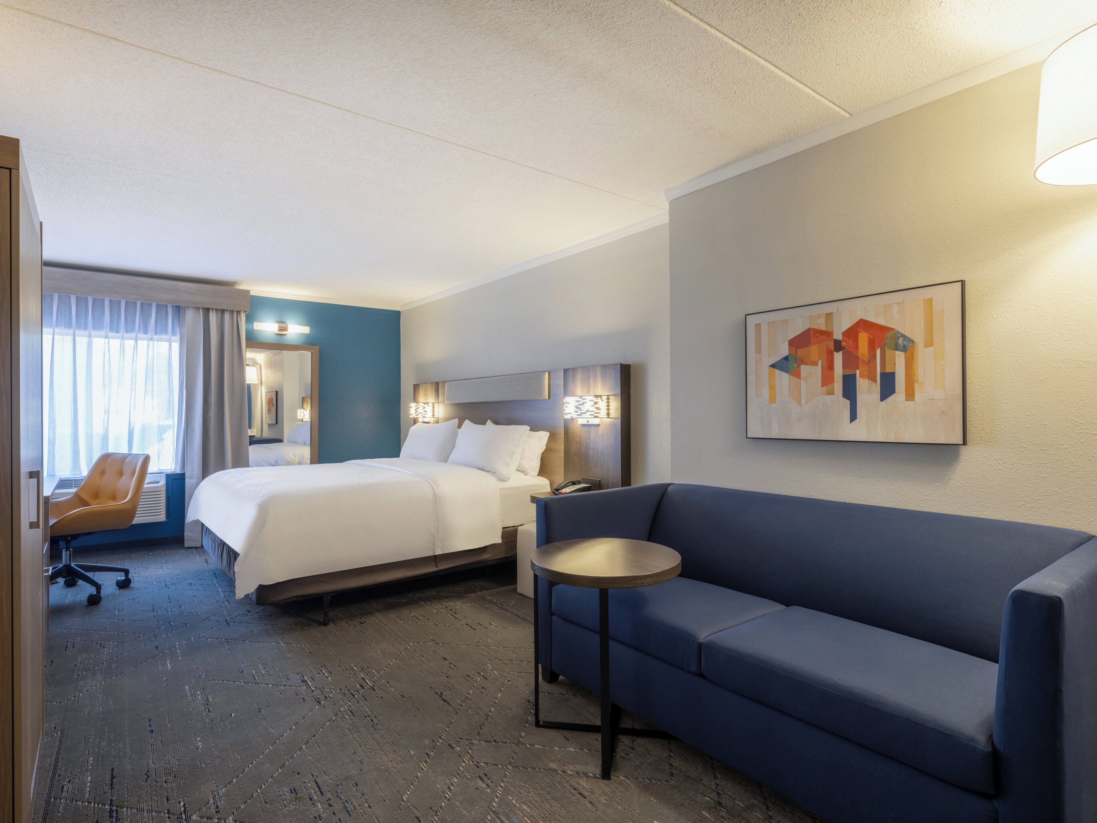 Quarto de hotel com uma cama, um sofá azul, uma mesinha redonda pequena, uma cadeira e um quadro na parede.