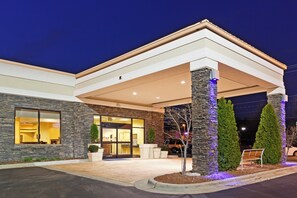A modern building with a stone facade, a covered entrance, and a well-maintained outdoor area with a bench and potted plants. - Holiday Inn Express & Suites Greensboro-(I-40 Wendover) by IHG (Greensboro)