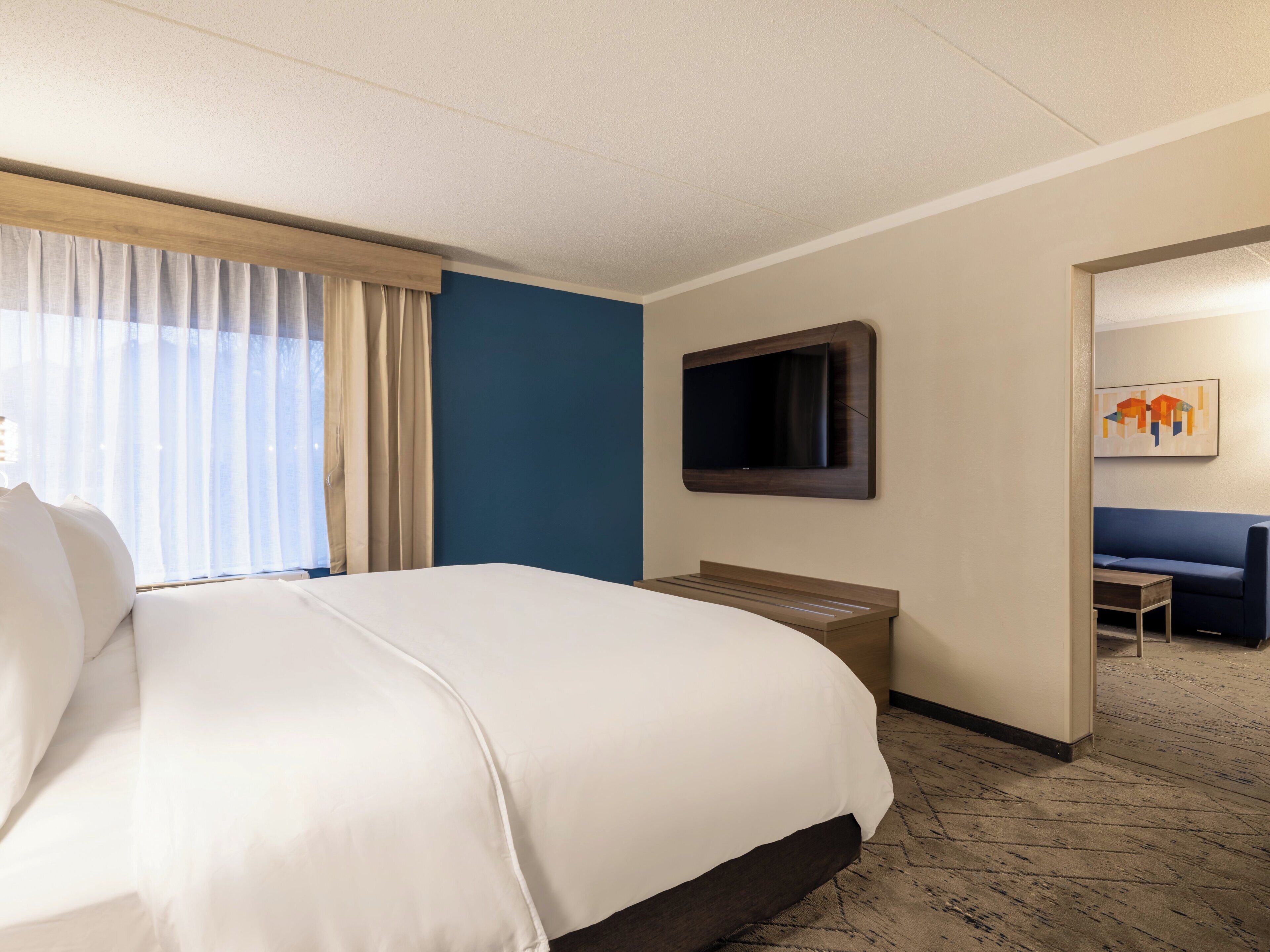 A hotel room with a large bed, a flat-screen TV mounted on the wall, a seating area with a sofa and a small table, and a painting on the wall.