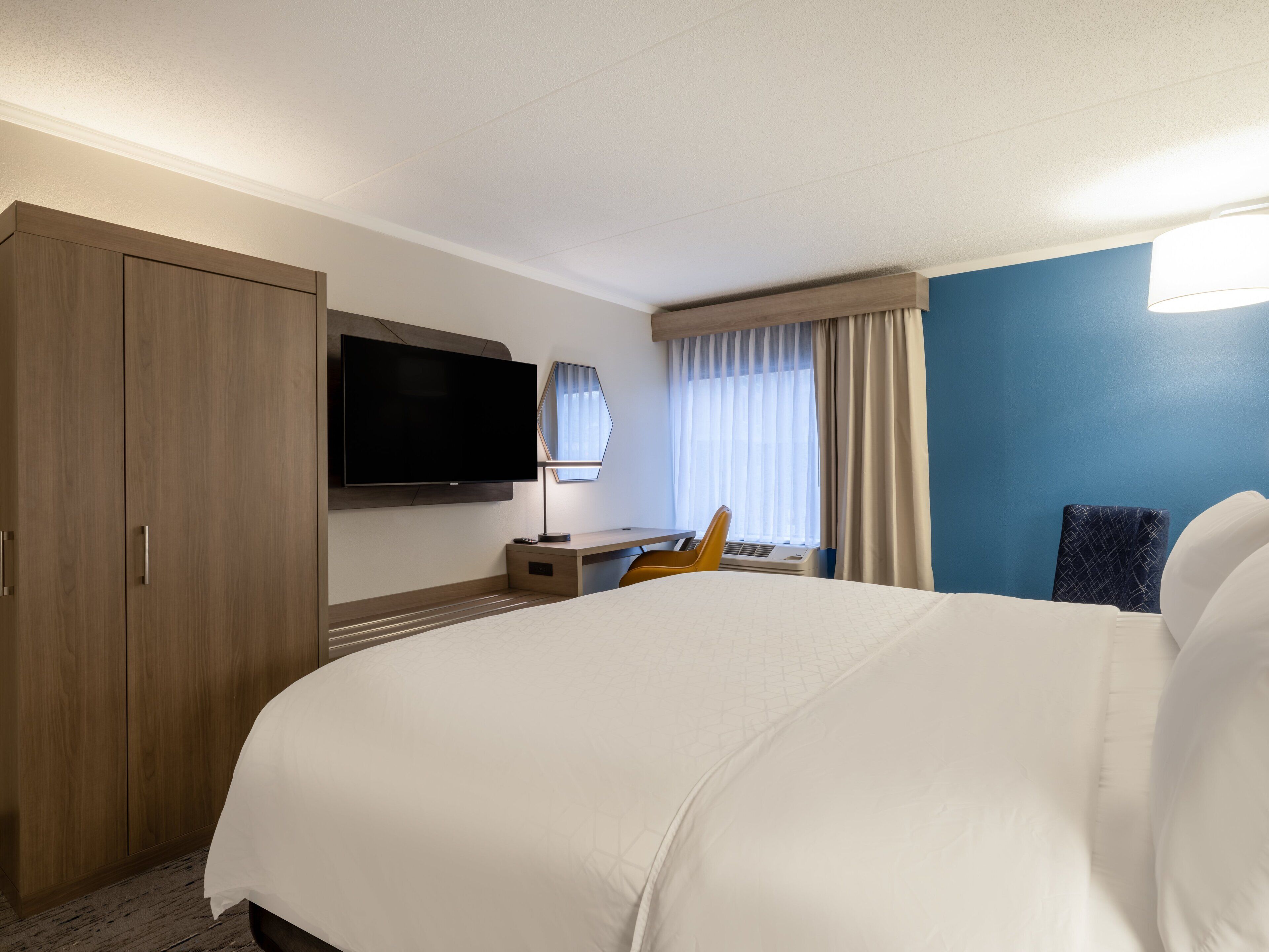 A hotel room with a large bed, a desk with a chair, a flat-screen TV mounted on the wall, and a window with curtains.