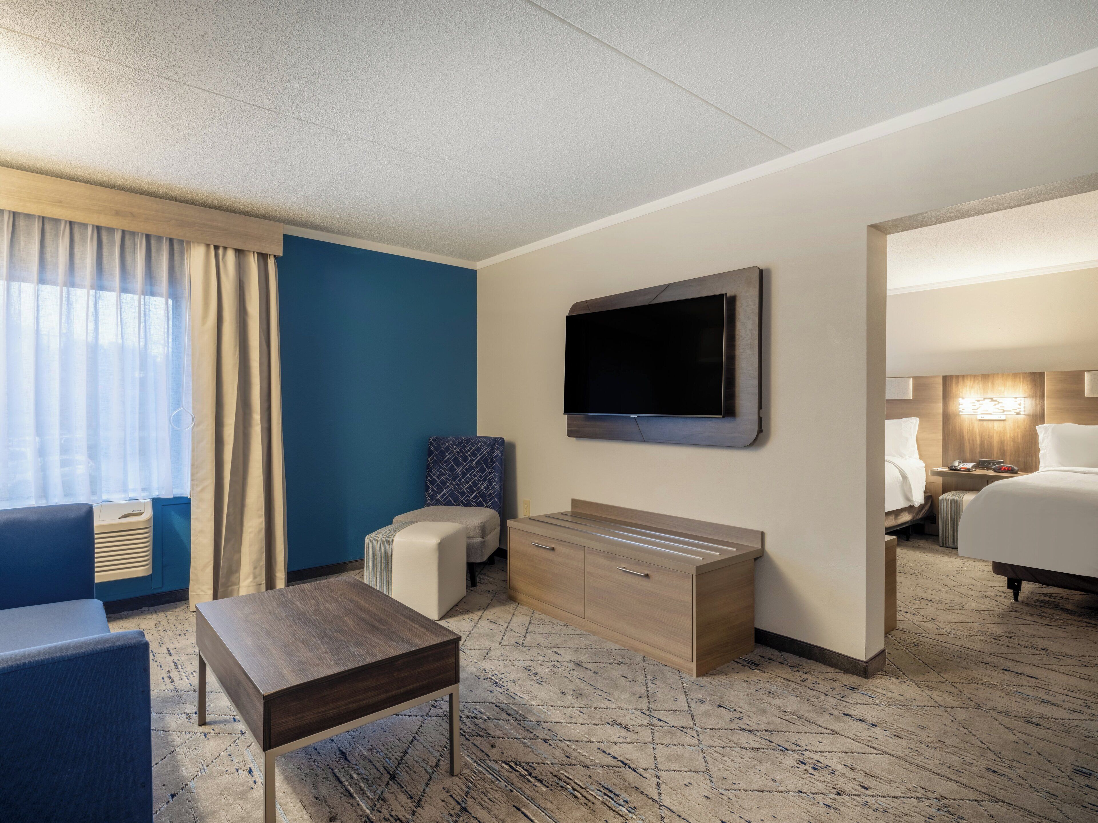Hotelkamer met flatscreen televisie aan de wand, een zithoek met een tafeltje en een bed met hoofdeinde.