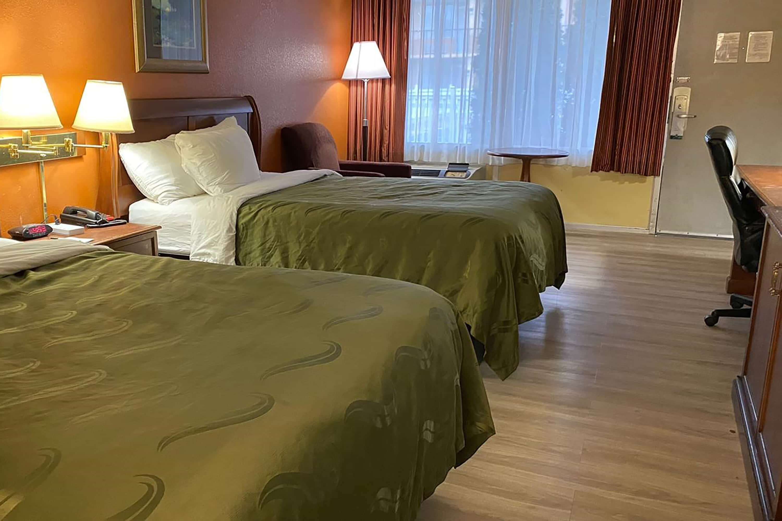 Habitación de hotel con dos camas, un escritorio, una silla, una lámpara y un ventanal con cortinas.