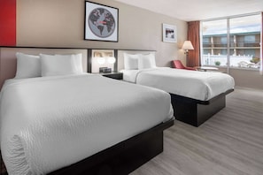 A hotel room with two beds, a nightstand with a clock, a lamp, and a large window with a view of buildings.