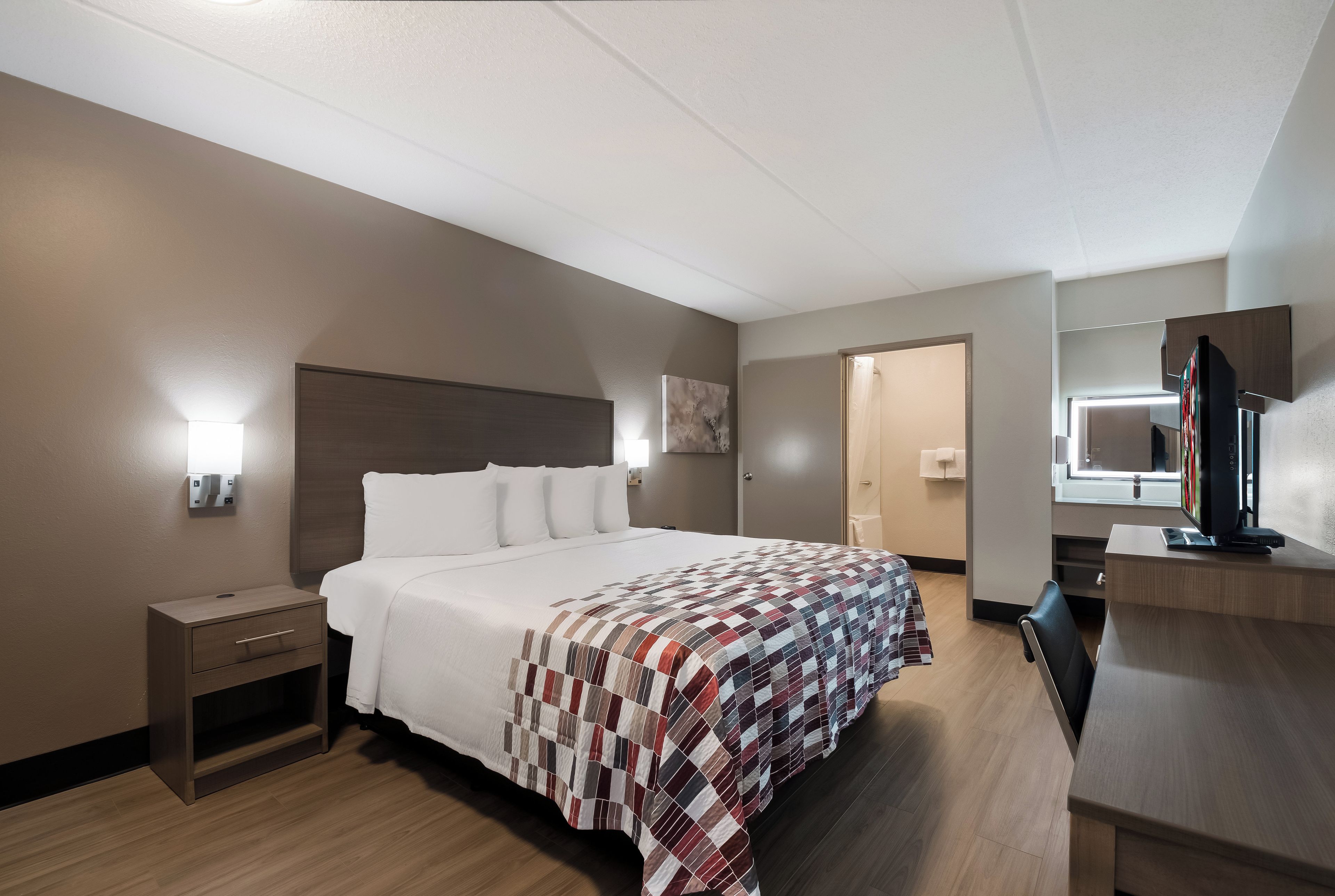 A hotel room with a bed, a nightstand, a desk with a computer, a mirror, and a shower area.