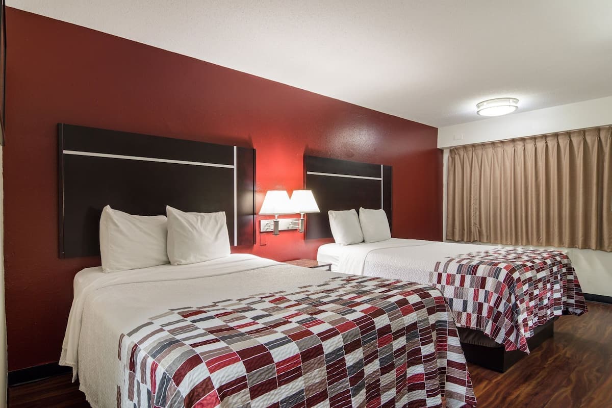 Hotellihuone, jossa on kaksi sänkyä, punainen tehosteseinä, kattovalaisin ja ikkuna, jossa on verhot.